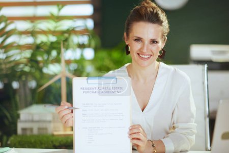 Photo for Sustainable real estate business. smiling elegant female realtor in modern green office in white blouse with clipboard and document. - Royalty Free Image