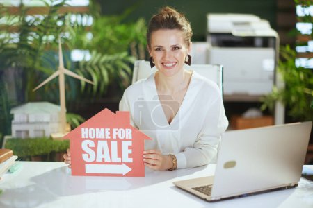 Photo for Sustainable real estate business. happy 40 years old business woman in modern green office in white blouse with home for sale sign and laptop. - Royalty Free Image