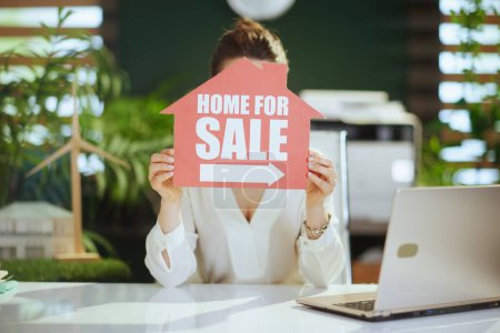 Téléchargez les photos : Entreprise immobilière durable. élégant 40 ans femme agent immobilier dans le bureau vert moderne en chemisier blanc avec signe de la maison à vendre et ordinateur portable. - en image libre de droit