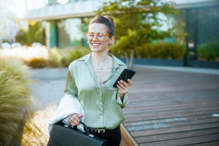 Téléchargez les photos : Femme d'affaires souriante moderne de 40 ans dans le quartier des affaires en chemisier vert et lunettes avec smartphone et mallette. - en image libre de droit