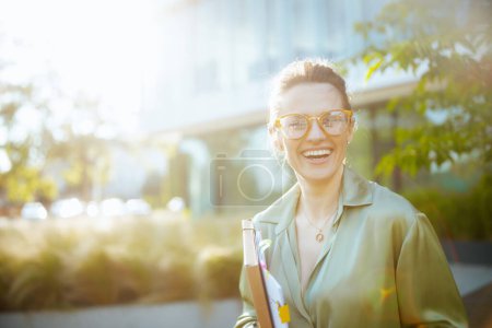 Foto de Retrato de la sonriente y moderna empleada de 40 años en el distrito de negocios en blusa verde y anteojos con documentos y carpeta. - Imagen libre de derechos