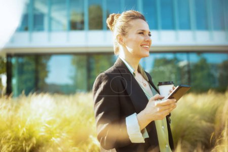 Foto de Sonriente trabajadora moderna en el distrito de negocios en chaqueta negra con taza de café usando teléfono inteligente. - Imagen libre de derechos