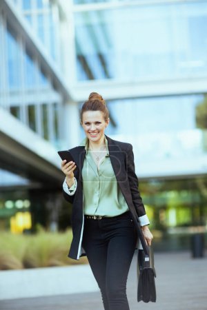 Foto de Sonriendo moderno 40 años de edad, mujer trabajadora cerca de edificio de oficinas en chaqueta negra con teléfono inteligente y maletín caminando. - Imagen libre de derechos