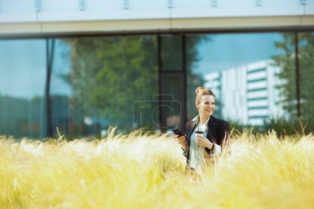 Photo for Smiling modern 40 years old woman employee near office building in black jacket with cup of coffee using smartphone. - Royalty Free Image