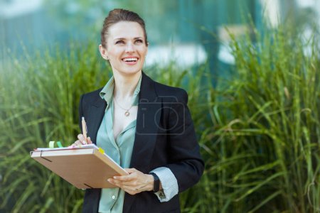Foto de Feliz mujer de negocios moderna de mediana edad cerca del edificio de oficinas en chaqueta negra con documentos y carpeta. - Imagen libre de derechos
