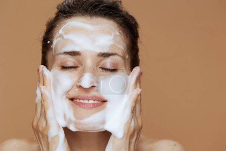Foto de Mujer joven y relajada con rostro espumoso lavado cara contra fondo beige. - Imagen libre de derechos