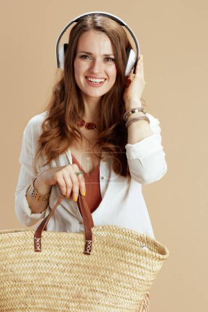 Beach vacation. smiling trendy female in white blouse and shorts isolated on beige background with straw bag listening to the music with headphones.
