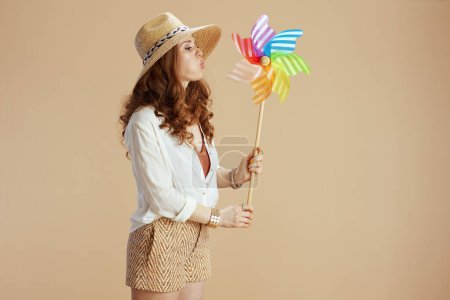 Photo for Beach vacation. modern middle aged housewife in white blouse and shorts isolated on beige background with windmill toy and summer hat. - Royalty Free Image
