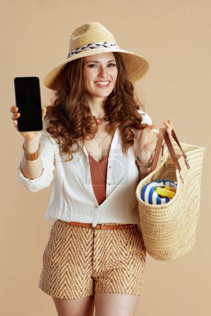 Téléchargez les photos : Vacances à la plage. heureuse femme moderne en chemisier blanc et short sur fond beige avec sac de paille et chapeau d'été montrant écran blanc smartphone. - en image libre de droit