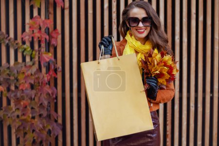Téléchargez les photos : Bonjour octobre. femme élégante souriante en trench-coat marron avec sac à provisions et feuilles jaune automne dans la ville. - en image libre de droit