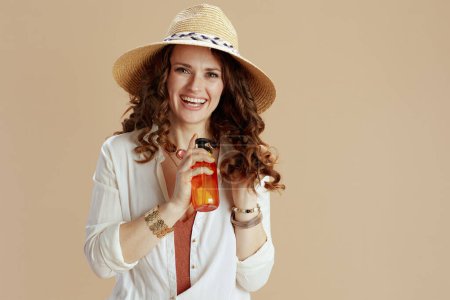 Photo for Beach vacation. smiling stylish 40 years old housewife in white blouse and shorts isolated on beige background with hair care product and straw hat. - Royalty Free Image