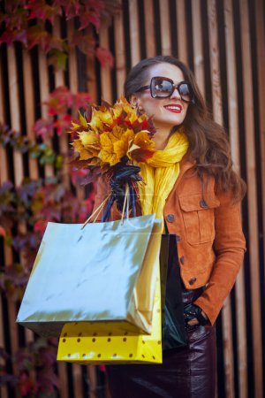 Foto de Hola noviembre. mujer moderna y relajada en gabardina marrón con bolsas de compras y hojas amarillas de otoño en la ciudad. - Imagen libre de derechos