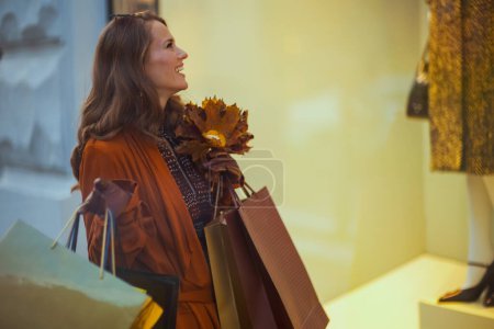 Foto de Hola octubre. elegante y sonriente mujer de 40 años de edad en gabardina marrón con bolsas de compras y hojas amarillas de otoño cerca de la tienda en la ciudad. - Imagen libre de derechos