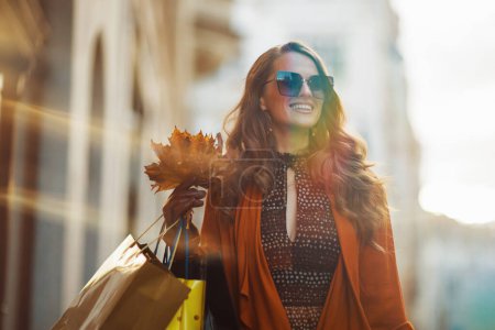 Photo for Hello october. smiling stylish female in orange trench coat with shopping bags and autumn yellow leaves in the city. - Royalty Free Image