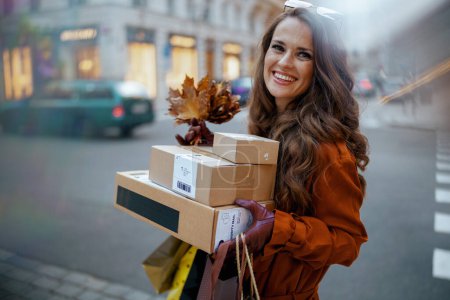 Photo for Hello november. happy trendy 40 years old woman in brown trench coat with parcels, shopping bags and autumn yellow leaves in the city. - Royalty Free Image