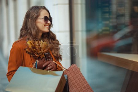 Foto de Hola noviembre. mujer con estilo feliz en gabardina marrón con bolsas de compras y hojas amarillas de otoño cerca de la tienda en la ciudad. - Imagen libre de derechos