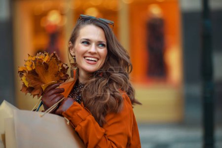 Foto de Hola otoño. mujer moderna sonriente en gabardina naranja con bolsas de compras y hojas amarillas de otoño cerca de la tienda en la ciudad. - Imagen libre de derechos