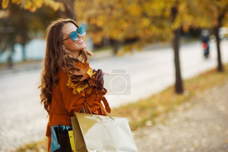 Foto de Hola septiembre. Elegante hembra sonriente en gabardina naranja con bolsas de compras y hojas amarillas otoñales en la ciudad. - Imagen libre de derechos