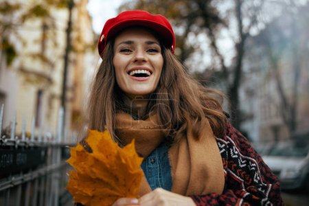 Foto de Hola otoño. Retrato de mujer de 40 años de moda feliz en sombrero rojo con hojas de otoño y bufanda en la ciudad. - Imagen libre de derechos