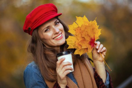 Photo for Hello autumn. Portrait of smiling trendy middle aged woman in jeans shirt and red hat with autumn leafs, scarf and coffee in the city park. - Royalty Free Image