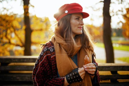 Foto de Hola otoño. mujer de mediana edad pensativa de moda en sombrero rojo con hoja de otoño, bufanda y guantes sentados en el banco en el parque de la ciudad. - Imagen libre de derechos