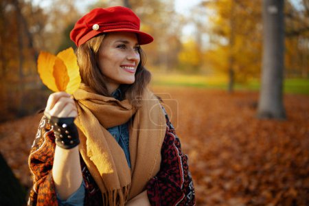 Foto de Hola otoño. mujer joven feliz en sombrero rojo con hojas de otoño, bufanda y guantes en el parque de la ciudad. - Imagen libre de derechos