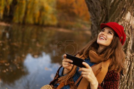 Foto de Hola otoño. mujer elegante relajado en sombrero rojo con bufanda usando teléfono inteligente cerca del árbol en el parque de la ciudad. - Imagen libre de derechos
