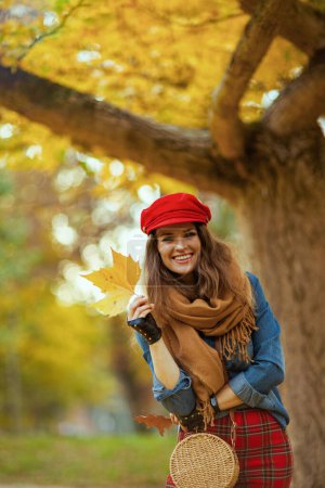 Téléchargez les photos : Bonjour l'automne. Portrait de femme élégante heureuse en jeans chemise et chapeau rouge avec feuille d'automne, écharpe, gants et sac près de l'arbre dans le parc de la ville. - en image libre de droit