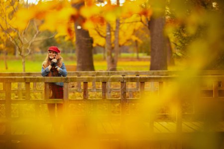 Foto de Hola otoño. mujer moderna sonriente en camisa de jeans y sombrero rojo con hoja de otoño y bufanda en el parque de la ciudad. - Imagen libre de derechos