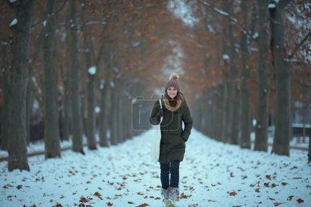 Foto de Retrato completo de mujer moderna feliz en abrigo verde y sombrero marrón al aire libre en el parque de la ciudad en invierno con sombrero de gorro. - Imagen libre de derechos