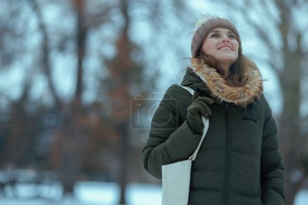 Foto de Mujer moderna feliz en abrigo verde y sombrero marrón al aire libre en el parque de la ciudad en invierno con manoplas y gorro sombrero. - Imagen libre de derechos
