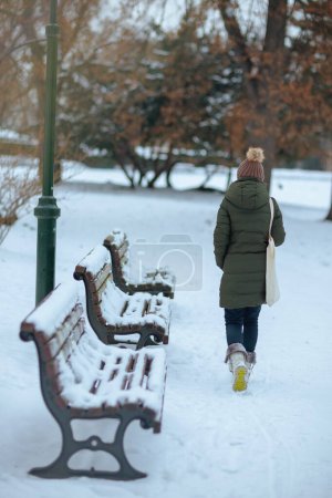 Foto de Visto desde atrás mujer moderna de 40 años de edad en abrigo verde y sombrero marrón al aire libre en el parque de la ciudad en invierno con gorro sombrero. - Imagen libre de derechos