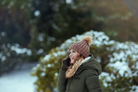 Photo for Smiling modern woman in green coat and brown hat outdoors in the city park in winter with mittens and beanie hat near snowy branches talking on a smartphone. - Royalty Free Image