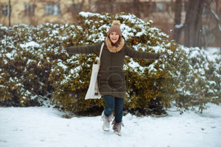 Téléchargez les photos : Portrait complet de femme moderne heureuse en manteau vert et chapeau brun à l'extérieur dans le parc de la ville en hiver avec des mitaines et un bonnet près des branches enneigées. - en image libre de droit