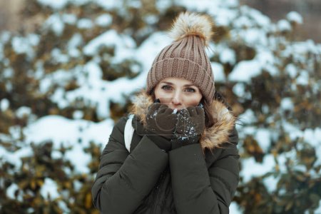Photo for Happy modern middle aged woman in green coat and brown hat outdoors in the city park in winter with mittens and beanie hat warming cold hands with breath near snowy branches. - Royalty Free Image