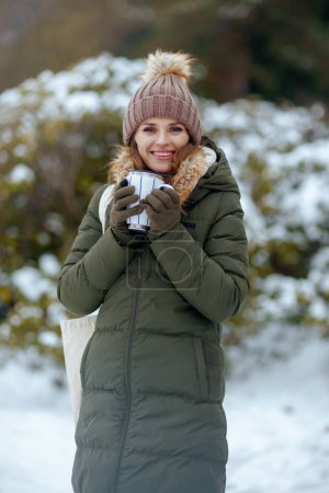 Foto de Sonriente mujer moderna en abrigo verde y sombrero marrón al aire libre en el parque de la ciudad en invierno con manoplas, taza de cacao caliente y sombrero de gorro. - Imagen libre de derechos