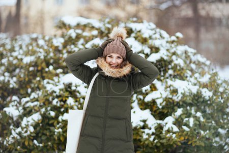 Foto de Mujer moderna feliz en abrigo verde y sombrero marrón al aire libre en el parque de la ciudad en invierno con manoplas y gorro sombrero cerca de ramas nevadas. - Imagen libre de derechos