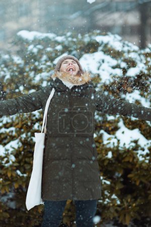 Foto de Sonriente mujer moderna en abrigo verde y sombrero marrón al aire libre en el parque de la ciudad en invierno con manoplas y gorro jugando con nieve cerca de ramas nevadas. - Imagen libre de derechos
