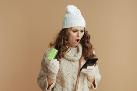 Photo for Hello winter. shocked elegant female in beige sweater, mittens and hat with smartphone and credit card buying online isolated on beige. - Royalty Free Image