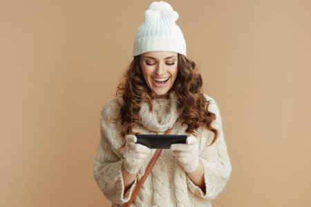 Photo for Hello winter. smiling trendy female in beige sweater, mittens and hat isolated on beige background using smartphone app. - Royalty Free Image