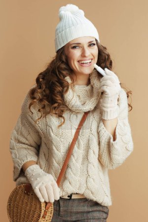 Téléchargez les photos : Bonjour l'hiver. Femme élégante et souriante en pull beige, mitaines et chapeau avec rouge à lèvres hygiénique isolé sur beige. - en image libre de droit