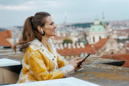 Foto de Mujer viajera solista de moda feliz en blusa amarilla y impermeable en Praga República Checa con auriculares usando teléfono inteligente contra panorama de la ciudad. - Imagen libre de derechos
