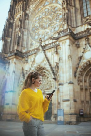 Téléchargez les photos : Heureuse voyageuse d'âge moyen à la mode en chemisier jaune à Prague République tchèque ayant une visite à pied, en utilisant un smartphone et la marche. - en image libre de droit