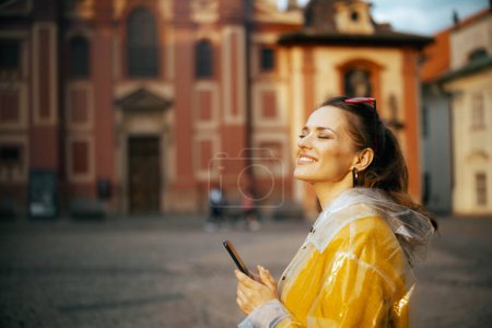 Téléchargez les photos : Femme voyageuse élégante souriante en chemisier jaune et imperméable à Prague République tchèque avec smartphone. - en image libre de droit