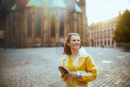 Photo for Smiling young traveller woman in yellow blouse and raincoat in Prague Czech Republic having walking tour, using smartphone and walking. - Royalty Free Image