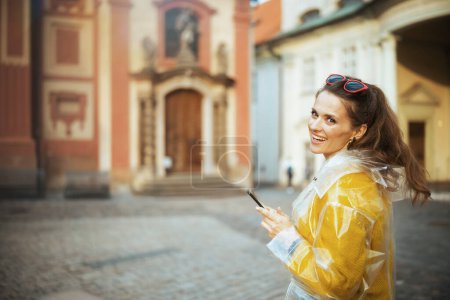 Téléchargez les photos : Femme souriante à la mode en chemisier jaune et imperméable à Prague République tchèque avec smartphone. - en image libre de droit