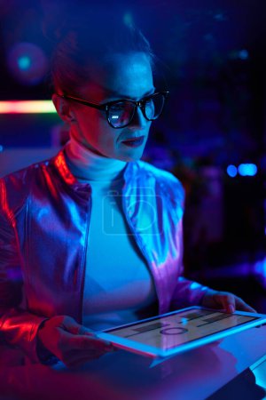 Photo for Neon metaverse futuristic concept. pensive modern business woman in glasses using tablet PC and exploring charts in modern office. - Royalty Free Image