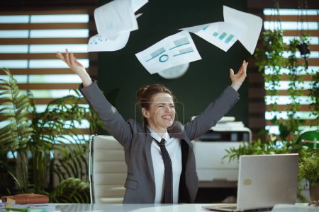Photo for New job. smiling modern middle aged woman worker in modern green office in grey business suit with laptop throwing documents. - Royalty Free Image