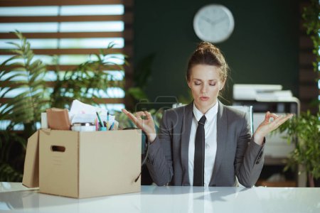 Photo for New job. stressed modern 40 years old woman employee in modern green office in grey business suit with personal belongings in cardboard box meditating. - Royalty Free Image
