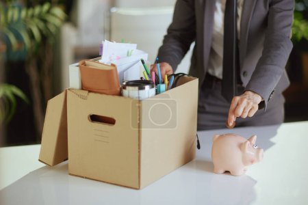 Photo for New job. Closeup on modern female worker in modern green office with personal belongings in cardboard box putting coin into piggy bank. - Royalty Free Image